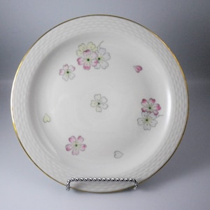 May include: A white ceramic plate with a gold rim and a textured border. The plate is decorated with pink and white floral designs and small heart shapes. The plate is displayed on a silver metal stand.