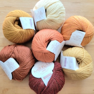 May include: Seven skeins of yarn in various warm colors, including mustard yellow, rust, and cream. Each skein has a label attached. The yarn is arranged on a light brown wooden surface, creating a visually appealing display for crafting projects.
