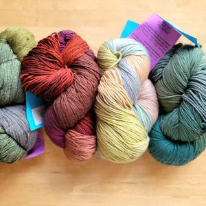 May include: A collection of colorful yarn skeins in shades of green, red, purple, and beige. Each skein is a different color and has a tag attached. The yarn is arranged on a light brown wooden surface.