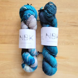 May include: Two skeins of colorful yarn in shades of blue, teal, and gray, with white labels that read "NBK". The yarn is arranged on a light brown wooden surface, showcasing the vibrant colors and textures of the fiber.
