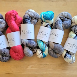May include: Six skeins of colorful yarn in shades of pink, gray, blue, and cream, each with a white label printed with "NBK". The yarn is arranged on a light brown wooden surface, showcasing the variety of colors and textures.