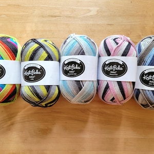 May include: Five colorful yarn skeins with white labels that read "Knit Picks Felici". The yarn colors include rainbow, black and yellow, blue and gray, pink and black, and blue and brown. The yarn is displayed on a wooden surface.
