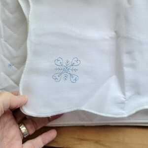 May include: White quilted fabric with a scalloped edge. A blue embroidered snowflake design is visible on the fabric. The fabric is folded, and a hand with a silver ring is holding the fabric.