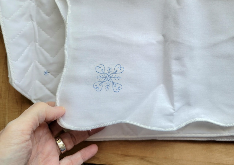 May include: White quilted fabric with a scalloped edge. A blue embroidered snowflake design is visible on the fabric. The fabric is folded, and a hand with a silver ring is holding the fabric.
