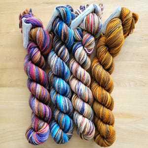 May include: Four skeins of colorful yarn in shades of purple, blue, brown, and gold are arranged on a light wood surface. Each skein is tightly wound and has a paper label attached. The yarn appears to be hand-dyed, with variations in color throughout.
