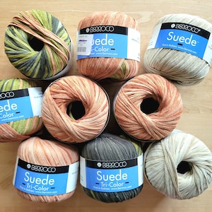 May include: A collection of yarn skeins in various colors, including green, brown, and gray. The yarn is labeled "Suede Tri-Color" by Berroco. The skeins are arranged on a light wooden surface.