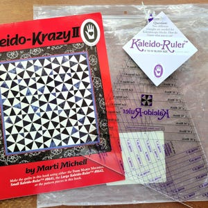 May include: A red instructional book titled Kaleido-Krazy II by Marti Michell featuring a black and white quilt pattern, paired with a clear plastic Kaleido-Ruler for 6 to 16 inch block sizes, packaged in a clear plastic bag with a white tag.