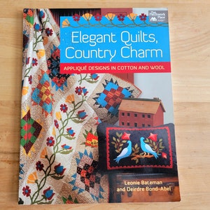 May include: A book titled "Elegant Quilts, Country Charm" with the subtitle "Appliqué Designs in Cotton and Wool." The cover features images of quilts and a small red house. The authors are Leonie Bateman and Deirdre Bond-Abel.