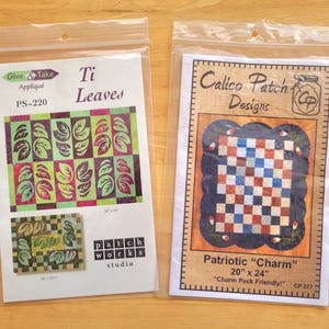 May include: Two packaged quilting patterns. The "Ti Leaves" pattern features a leaf design in green, purple, and pink. The "Patriotic Charm" pattern displays a red, white, and blue checkerboard design, measuring 20" x 24".