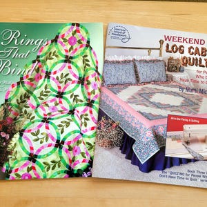 May include: Two open books on a wooden surface. The left book cover reads "Rings That Bind" with a quilt design. The right book cover says "Weekend Log Cabin Quilts" with a bed quilt design. Both books are about quilting.