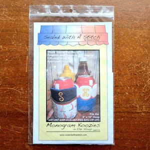 May include: A clear plastic package displays a printed image of two beverage can koozies, a mustard bottle, and a ketchup bottle. The koozies are decorated with nautical and monogram designs. Text on the package reads "Sealed With A Stitch" and "Monogram Koozies in the Hoop."