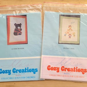 May include: Two sealed plastic bags containing children's quilt kits. One features a bear with a red bow, the other shows a stack of bears. The bags have a light blue and pink label with the text "Cozy Creations" and "Collectible Quilts for Children."