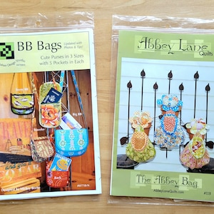 May include: Two sewing pattern booklets in clear plastic sleeves. The left booklet is titled BB Bags and shows various small purses with floral and geometric patterns. The right booklet is titled The Abbey Bag and displays three fabric bags.