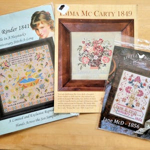 May include: Three framed cross-stitch samplers. The top one is labeled "Emma Mc Carty 1849" with a floral design. The left one is "Sarah Rinder 1841" with a portrait and a detailed needlework design. The third is "Jane McD - 1856".