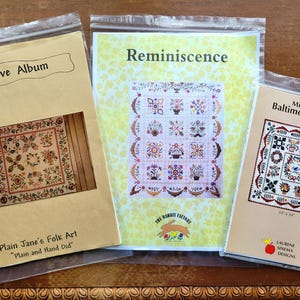 May include: Three quilt pattern booklets in clear plastic sleeves. The left booklet is titled Primitive Album by Plain Jane's Folk Art. The center booklet is titled Reminiscence by The Rabbit Factory. The right booklet is Miniature Baltimore Album by Laurene Sinema Designs, noting 34" x 34" dimensions.