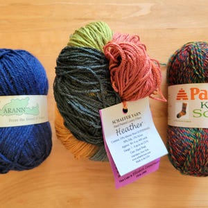 May include: Three skeins of yarn in various colors sit on a wooden surface. A blue skein with a white label that reads "Arann" is on the left. In the center is a multi-colored skein with a tag that reads "Heather". On the right is a skein labeled "Paton Kroy Socks FX".