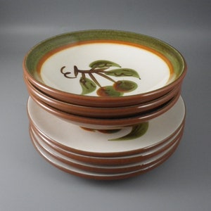 May include: A stack of six ceramic plates and bowls featuring a cream-colored center with a hand-painted green and brown cherry motif. The rims are finished with a wide band of olive green and a thin orange stripe, set against a solid brown exterior.