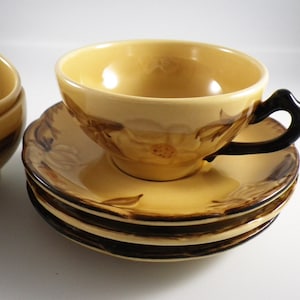 May include: A set of vintage teacups and saucers. The cups and saucers are a light yellow with brown floral designs. The cup has a dark brown handle. The saucers are stacked, and the set is on a white surface.