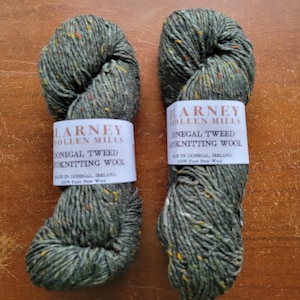 May include: Two skeins of green Donegal Tweed knitting wool from Blarney Woollen Mills. The yarn has flecks of yellow and orange. Each skein is wrapped with a white label that includes the brand name and product information. The yarn is 100% pure new wool.