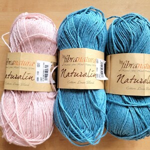 May include: Three skeins of yarn in shades of pink and teal blue, labeled "Naturalin Cotton Linen Blend" by "fibranatura." The yarn is wrapped with a brown paper band. The skeins are arranged on a light brown wooden surface.