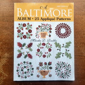 May include: A softcover book titled A Baltimore Album: 25 Appliqué Patterns by Marsha D. Radtke. The cover features a white background with nine distinct floral and wreath appliqué designs in shades of red, green, and brown, published by AGS.