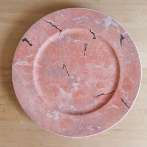 May include: A round, decorative plate with a marble-like design. The plate features a blend of pink, white, and gray hues, with black vein-like accents. The plate has a white rim and is suitable for serving food.