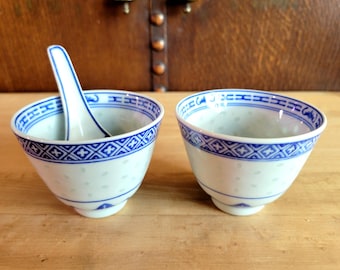 May include: Two white porcelain rice bowls with blue geometric patterns and a matching spoon. The bowls have a delicate dotted texture and a blue patterned rim. The spoon rests in one bowl.