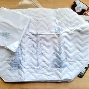 May include: White quilted placemat with blue embroidered winter scene, featuring snowmen and a tree. Includes a matching napkin with a snowflake design. The placemat has a zig-zag pattern.
