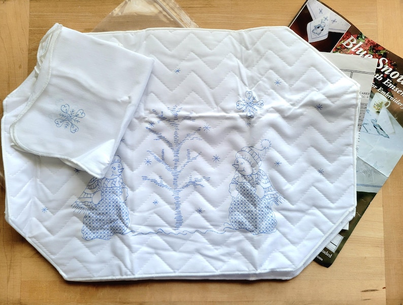 May include: White quilted placemat with blue embroidered winter scene, featuring snowmen and a tree. Includes a matching napkin with a snowflake design. The placemat has a zig-zag pattern.