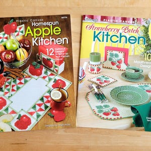 May include: Two craft pattern books on a wooden surface. The left book is titled "Homespun Apple Kitchen" with apple-themed designs. The right book is "Strawberry Patch Kitchen" with strawberry designs. Both books feature colorful kitchen decor.