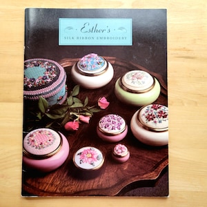 May include: A collection of decorative, lidded trinket boxes with floral designs, displayed on a wooden tray. The boxes vary in size, shape, and color, including pink, white, and green. The image also features a book titled "Esther's Silk Ribbon Embroidery."