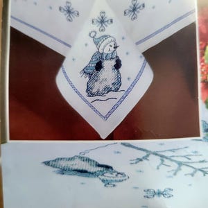 May include: White tablecloth with a blue embroidered snowman design. The snowman wears a blue hat and scarf. The tablecloth has a blue border and snowflake accents, perfect for winter holiday decor.