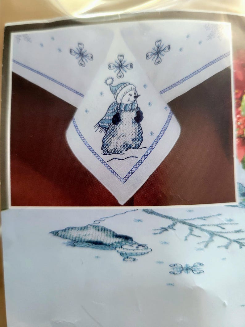 May include: White tablecloth with a blue embroidered snowman design. The snowman wears a blue hat and scarf. The tablecloth has a blue border and snowflake accents, perfect for winter holiday decor.