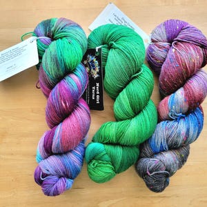 May include: Three skeins of colorful yarn in shades of green, purple, and blue, with attached tags. The yarn is arranged on a light brown wooden surface. The yarn is likely for knitting or crocheting.