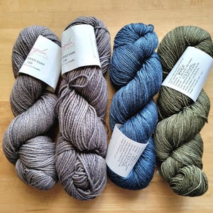 May include: Four skeins of dyed yarn in shades of gray, blue, and green, with white labels. The yarn is arranged on a light brown wooden surface. The labels have text, but it is too small to read.