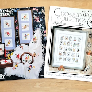 May include: Two cross-stitch pattern books. One titled "Nestling Sampler" features framed bird designs and a floral background. The other, "Crossed Wing Collection Alphabirds," showcases an alphabet with bird illustrations. Both are on a wooden surface.