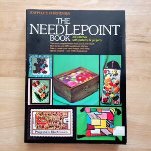 May include: The Needlepoint Book by Jo Ippolito Christensen. The book cover features needlepoint examples, including a teddy bear, flowers, and a box of flowers. The book promises over 300 stitches and 1000 illustrations.