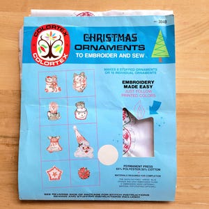 May include: A blue package of Colortex Christmas ornaments to embroider and sew. The package features illustrations of various Christmas ornaments, including a snowman, Santa Claus, and a star. The text "Embroidery Made Easy" is also visible.