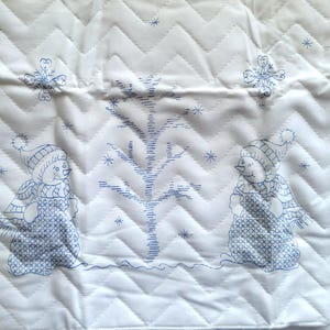 May include: White quilted fabric with a blue stitched design featuring two snowmen, a tree, and snowflakes. The fabric has a chevron pattern and is likely a decorative item for the winter season.