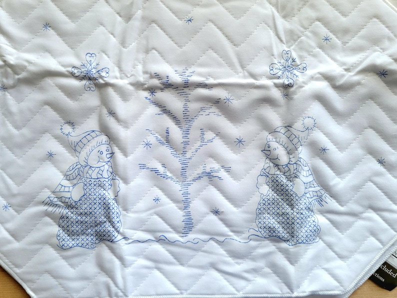 May include: White quilted fabric with a blue stitched design featuring two snowmen, a tree, and snowflakes. The fabric has a chevron pattern and is likely a decorative item for the winter season.