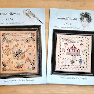 May include: Two framed samplers, "Anne Thomas 1854" and "Sarah Howarth 1835", feature detailed cross-stitch designs. The Anne Thomas sampler showcases a bird and floral motifs. The Sarah Howarth sampler depicts a house and figures. Both are from "Hands Across the Sea Samplers."