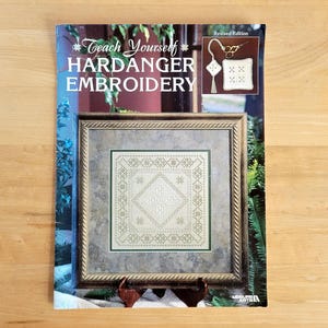 May include: A book titled "Teach Yourself Hardanger Embroidery" with a framed embroidery sample. The cover features the title in white and a small image of a finished project. The framed embroidery is a square design with a geometric pattern.