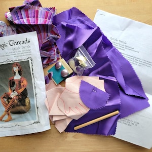 May include: A craft kit for a 24-inch cloth doll titled Razzle Dazzle by Magic Threads. Includes printed instruction sheets, purple fabric, light pink fabric pieces with hand outlines, a small bag of gold-toned charms, two purple buttons, and a wooden dowel.