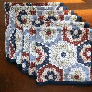 May include: A set of six quilted placemats with a patchwork design. The placemats feature a repeating pattern of hexagons in shades of blue, red, and white, with a dark blue border. The placemats are arranged in a fan shape.