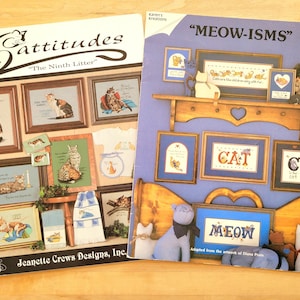 May include: Two books with cat-themed cross-stitch patterns. The left book is titled "Cattitudes" and features framed cat images. The right book, "MEOW-ISMS," showcases framed cat art and cat-themed decor. The books are on a wooden surface.