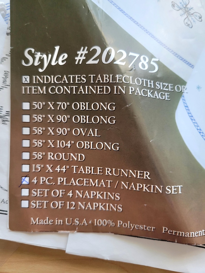 May include: A product label with the text "Style #202785" and a list of tablecloth sizes, including 50" x 70" oblong, 58" x 90" oblong, and 58" x 90" oval. The label also indicates a 4-piece placemat/napkin set.