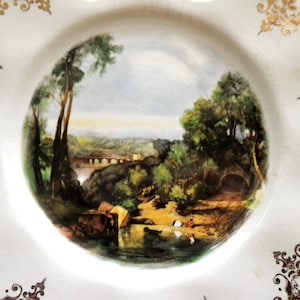 May include: White octagonal plate with a central landscape scene featuring a river, trees, and a bridge. The plate has gold filigree designs around the edges. The landscape scene is painted in shades of green, brown, and blue.