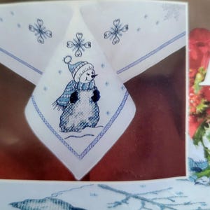 May include: White tablecloth with a blue embroidered snowman design. The snowman wears a blue hat and scarf. The tablecloth also features embroidered snowflakes and a blue border. A festive winter-themed item.