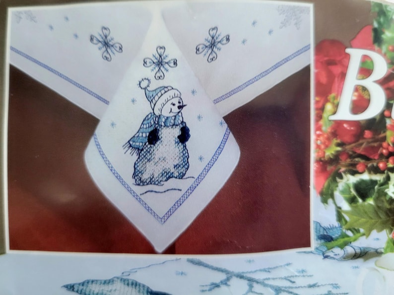 May include: White tablecloth with a blue embroidered snowman design. The snowman wears a blue hat and scarf. The tablecloth also features embroidered snowflakes and a blue border. A festive winter-themed item.