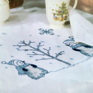 May include: White tablecloth with embroidered winter scene featuring snowmen, snowflakes, and a bare tree in blue and white. A sugar bowl and a mug with a Christmas tree design are in the background.